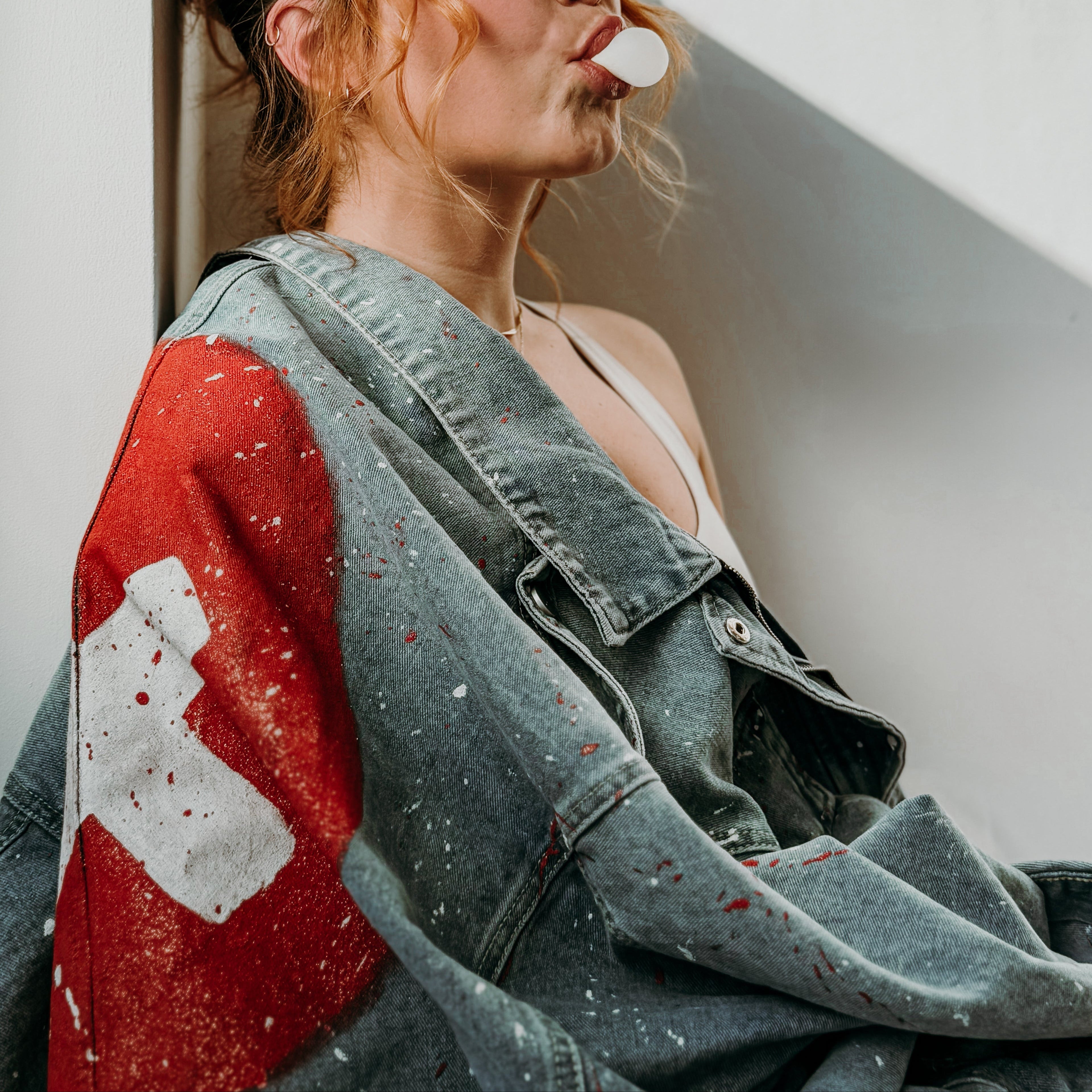 Hand-painted denim jacket with bold red and white design, worn by a woman leaning against a wall – RepeteArt wearable art.
